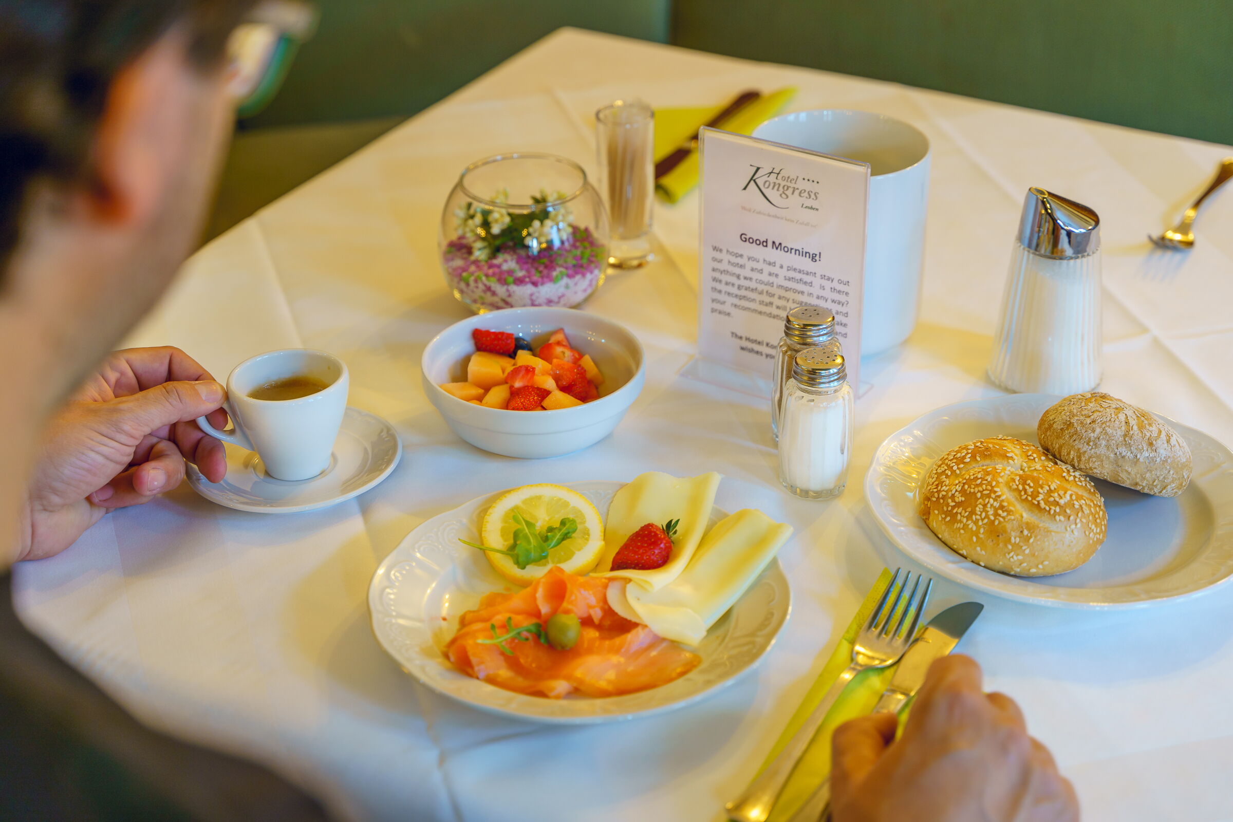 Frühstückstisch mit einem Teller Lachs und Käse, Brötchen, einem Obstsalat und eines Tasse Kaffee