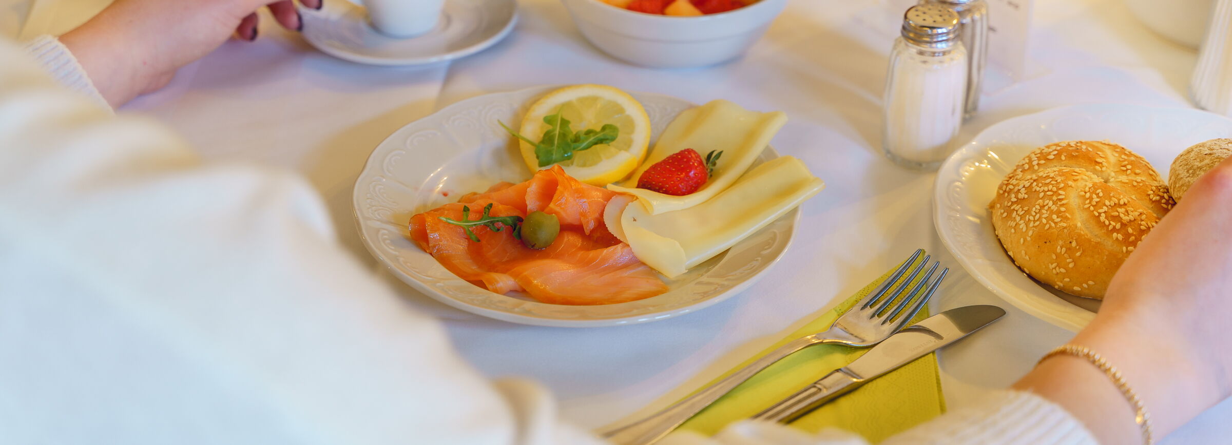 Frühstückstisch mit einem Teller Lachs und Käse, Brötchen und einem Obstsalat